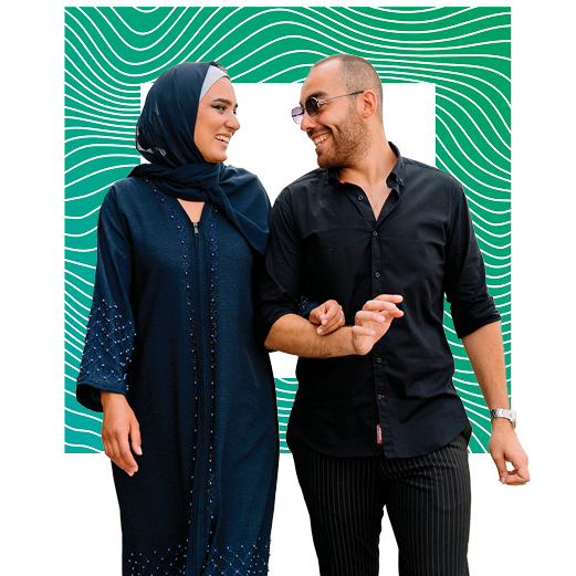 One person in a blue dress and hijab linking arms and smiling at another person in a dark button down and sunglasses smiling back.