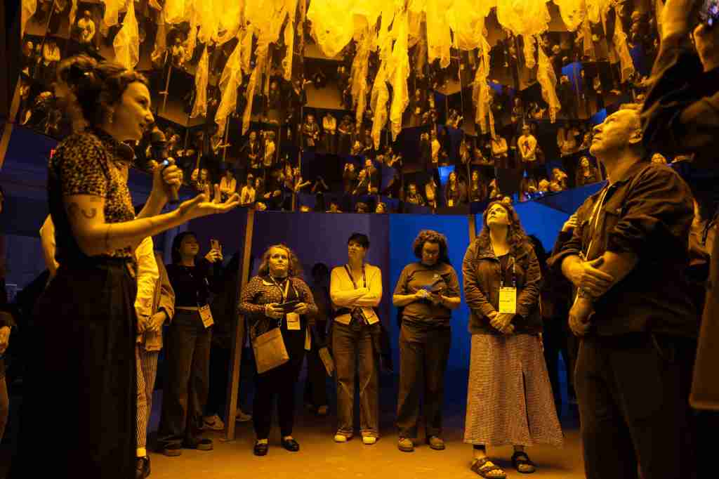 A group of people receive a museum tour. They look up at yellow lit mirror installation while someone speaks into a microphone.