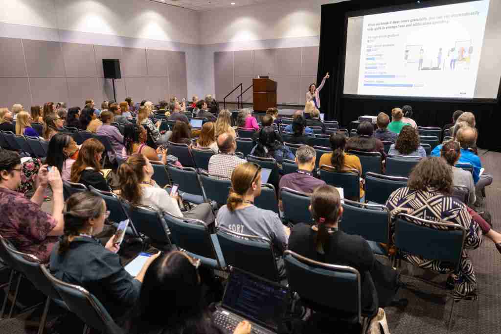 A full session room with a speaker pointing up at a large screen