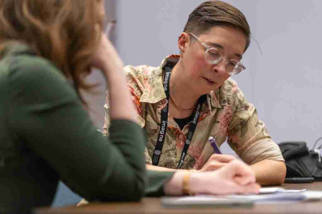 A person looks down at a sheet of paper while writing. Another person watches.