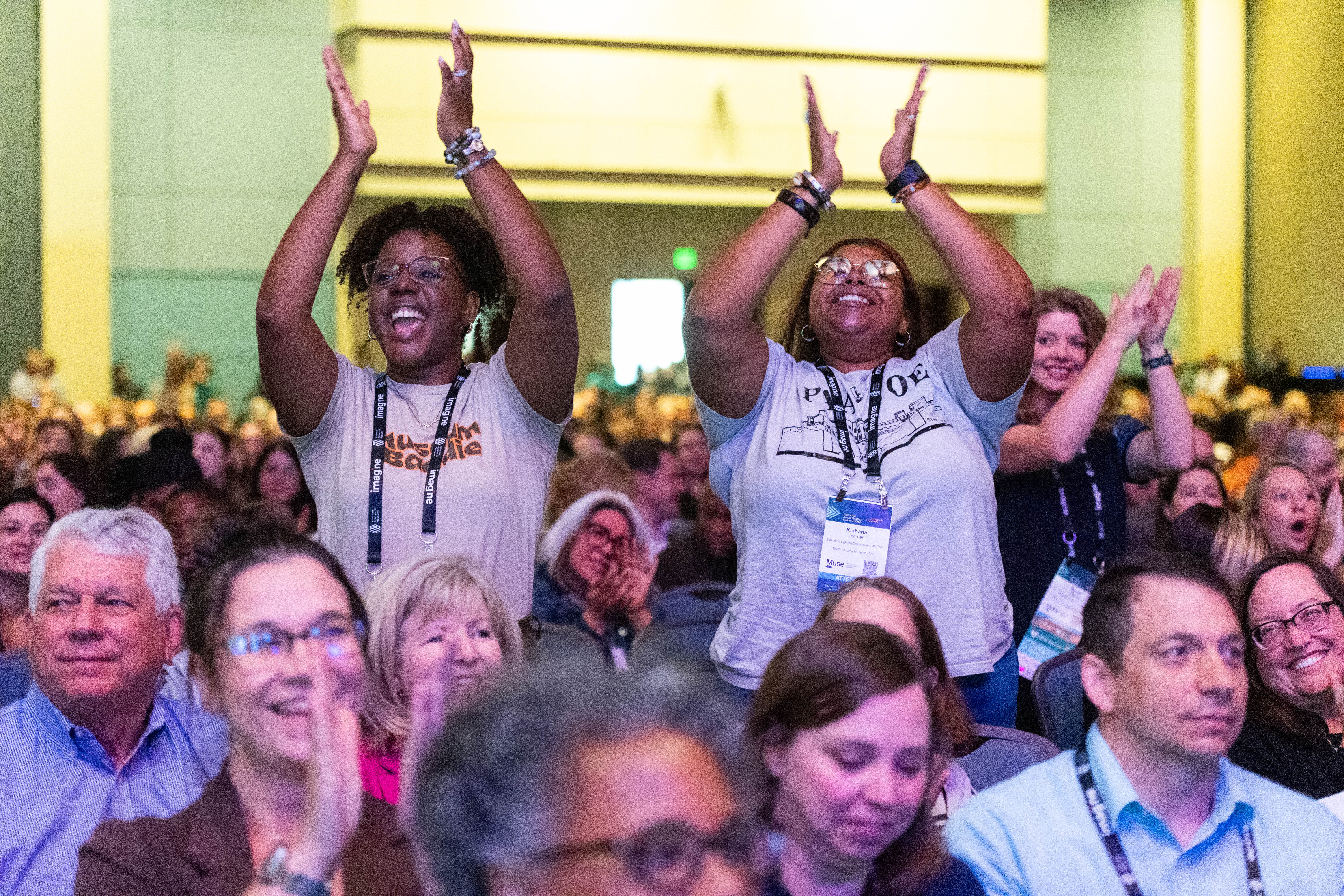 A crowd of attendees clapping during the general session with two attendees standing and emphatically clapping their hands in the foreground