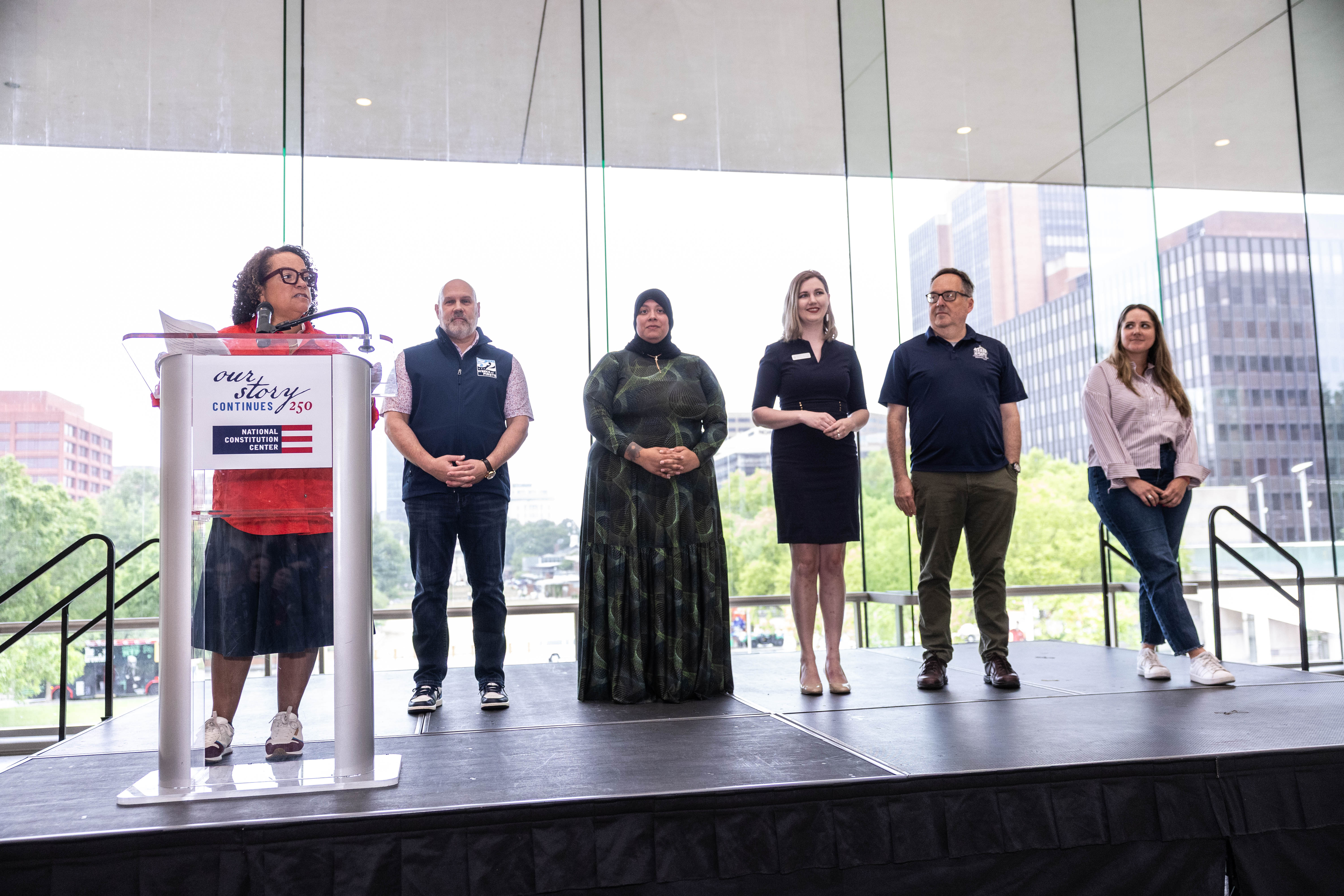 A group of leaders from Philadelphia's museums stand on stage while AAM President & CEO Marilyn Jackson speaks at a podium