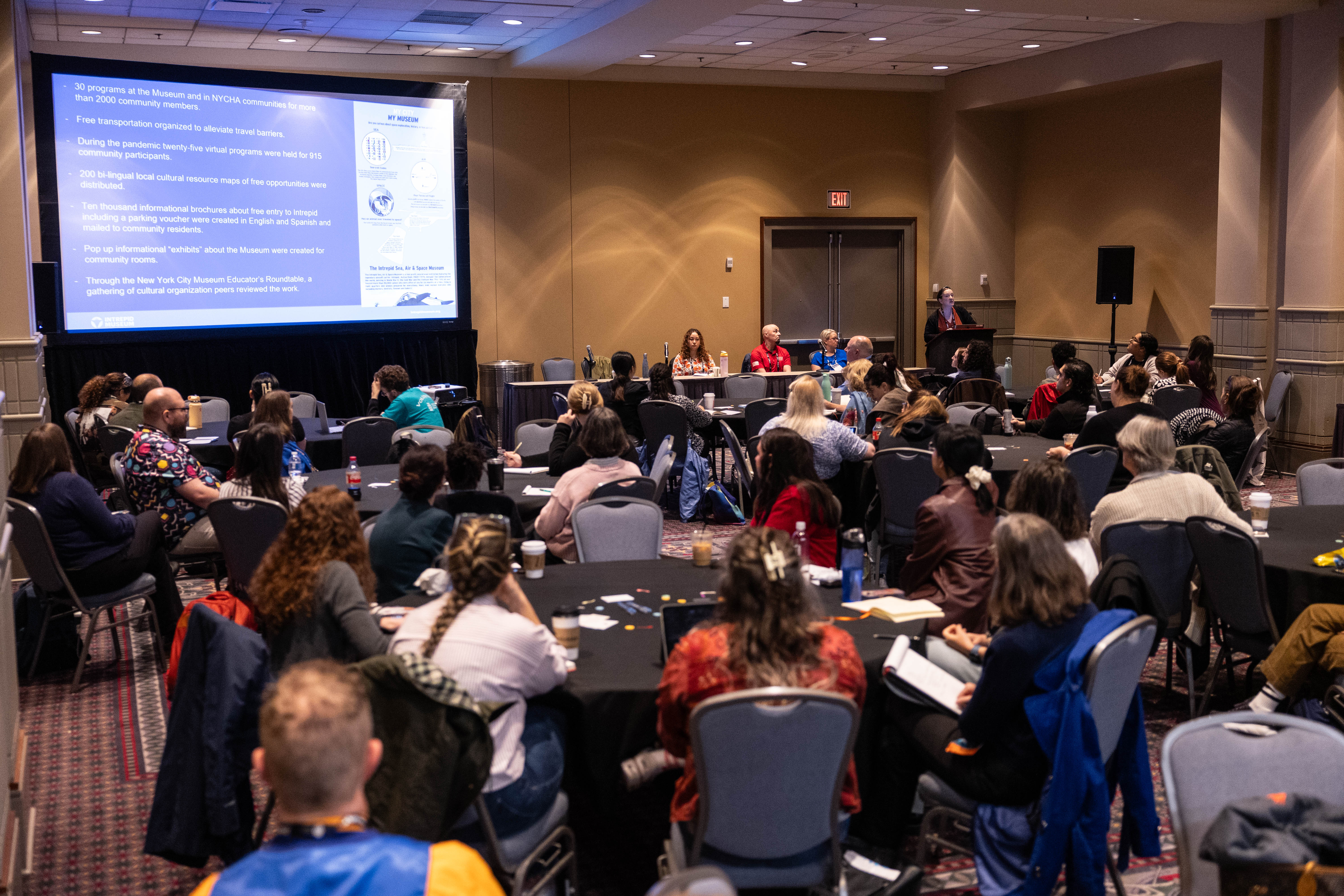 A full room of round tables during a panel presentation